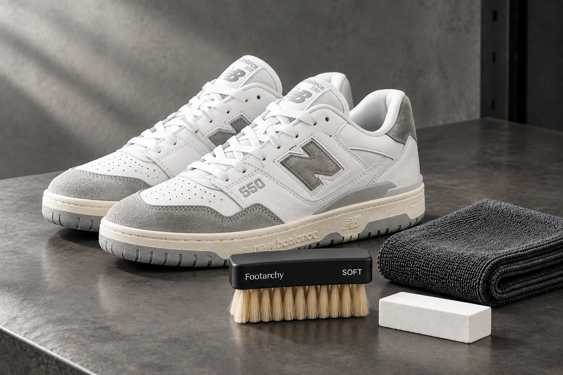 New Balance 550 blancas y grises junto a cepillo suave negro Footarchy, microfibra y borrador blanco sobre mesa industrial.